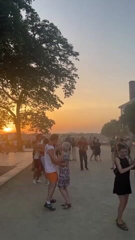 A group of people are dancing in an outdoor setting during sunset. The sky is a gradient of orange and yellow, with the sun partially visible behind a large tree on the left side of the frame. The ground is a light-colored gravel or dirt path. In the foreground, a young man and an older woman are dancing closely together, the man in a white tank top and orange shorts, the woman in a floral dress. To their right, another couple is dancing, the woman in a black dress and the man in a dark shirt. Further back, a person is playing an accordion, and other people are visible milling about and dancing. The overall atmosphere appears festive and relaxed.