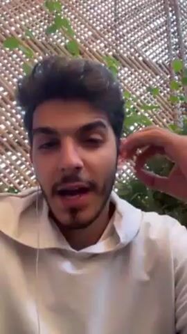 A young man with dark hair and a short beard speaks to the camera. He is wearing a white hoodie and has earbuds in. He gestures with his right hand. Behind him is a woven bamboo screen with green vines. He says “only took like three hours for editing this video.”