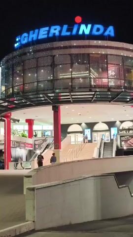 The video shows the exterior of a building at night. A large sign with blue lettering that reads "GHERLINDA" is visible at the top of the building, with a red circle above the "I". The building has a curved glass facade decorated with strings of small, twinkling lights. Below the sign, there is an entrance area with a curved canopy supported by red pillars. Several sets of stairs and escalators lead up to the entrance. There are some people visible in the video. Two individuals are standing on a walkway in front of the stairs. Another person appears to be walking down an escalator on the left side of the frame. The overall lighting suggests it is nighttime, and the environment appears to be an urban or commercial area.