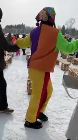The video depicts a winter scene with people participating in a festive activity. The ground is covered in snow. The participants are holding hands and walking over a series of wooden pallets placed on the snow.  

Among the participants, there are individuals in colorful costumes, possibly related to a cultural celebration or performance. They wear outfits that include bright colors like yellow, orange, and purple. 

Other people are dressed in typical winter clothing such as coats, hats, and scarves. The weather seems cold but sunny.

The overall atmosphere is lively and communal, suggesting a public event or gathering. In the background, there are buildings and trees, indicating an outdoor location, possibly a park or open space.