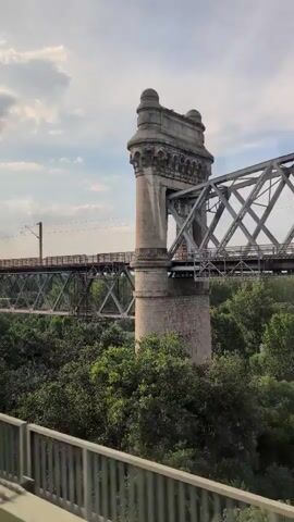 The video shows a view from a moving vehicle, likely a train or car, passing by a large bridge. The bridge appears to be a railway bridge with a metal truss structure supported by concrete piers. There are overhead power lines for an electric train visible above the bridge. The bridge spans over a dense area of green trees. On the right side of the frame, a section of a decorative stone tower, possibly part of the bridge's original architecture, is visible. The sky is partly cloudy, suggesting daytime. The camera angle is slightly downward, looking towards the bridge.