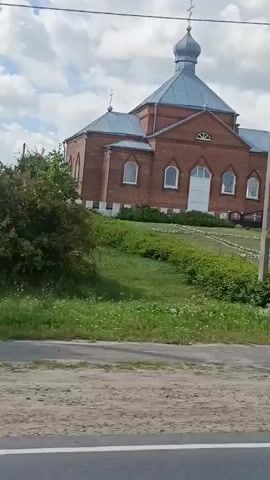 A moving shot captures the exterior of a red brick church with a blue dome. The church sits on a grassy hill, with a pathway leading up to its entrance. A utility pole stands in the foreground, partially obscuring the view of the church. The sky is partly cloudy. The camera appears to be inside or next to a vehicle, indicated by the partial view of a car in the bottom left of the frame and the movement of the scene. The overall impression is of passing by a religious building on a sunny day.