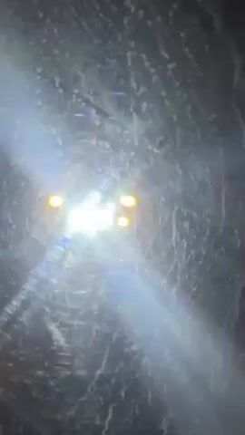 The video shows a bright light coming from a vehicle moving toward the camera. It is dark, with visible debris on the windshield. As the vehicle gets closer, a yellow car is seen from the side.