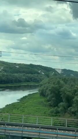 The video shows a view from a moving train window. The landscape consists of a wide river, lush green trees lining the riverbanks, and rolling hills in the distance. There are some white cliffs visible on the hills. The sky is cloudy. A train bridge is also visible in the foreground.