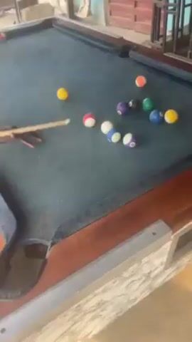 A person is playing pool on a pool table. There are several balls scattered across the table, including a black ball and several striped and solid-colored balls. The person is holding a cue stick and has just struck the cue ball. The cue ball is near the bottom center of the table, and the person's hand is resting on the table near it. The table has a wooden frame and a blue felt surface. A pocket is visible at the bottom left corner of the table, lined with a black material that says "PREMIER". The room where the pool table is located appears to be an outdoor or semi-outdoor setting, with visible wooden structures and sunlight shining through.