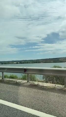 The video captures a wide landscape view from a moving vehicle, likely a car, looking across a large body of water towards a distant landmass. In the foreground, a metal guardrail stretches horizontally across the lower third of the frame, with reflective red markers visible on its surface. Below the guardrail, a gravelly roadside with sparse dry grass and small bushes is visible.

Beyond the guardrail, a vast expanse of calm, greyish-blue water dominates the middle ground. On the far bank of the water, a town or city skyline is visible, featuring various buildings, including what appear to be industrial structures or factories, and some taller buildings. The distant land is covered with greenery.

The sky is partly cloudy with large white and grey clouds, suggesting a bright daytime setting. A faint white contrail from an aircraft can be seen briefly in the upper left of the sky. The camera's perspective shifts slightly from left to right as the vehicle moves forward, offering a panoramic sweep of the scenery. The overall impression is one of traveling alongside a significant waterway. The provided context indicates this is near Drobeta-Turnu Severin, Romania, suggesting the body of water is the Danube River.