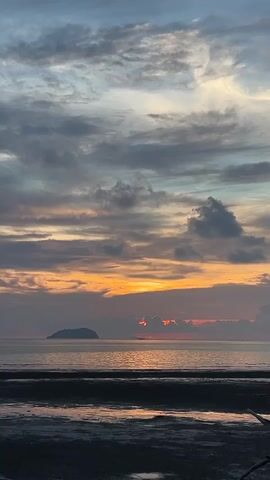A vertical, still shot captures a serene coastal sunset. The sky is a canvas of muted blues, grays, and oranges, with streaks of yellow and pink dominating the horizon where the sun is setting. Wispy clouds are scattered across the sky, some tinged with the warm hues of the sunset. Below the sky, a calm sea stretches to the horizon, reflecting the colors of the sky. In the distance, a small, dark island is silhouetted against the bright horizon. The foreground shows a dark, sandy or muddy shoreline, with a slight shimmer of water reflecting the sky. The overall atmosphere is tranquil and peaceful.