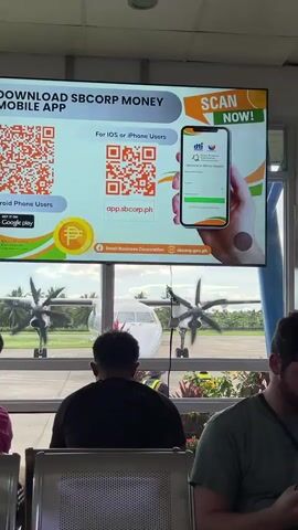 From inside an airport terminal, a monitor displays an advertisement for the SBCorp Money Mobile App. In the foreground, are several people seated, facing away from the camera. Through the terminal window, the propellers of a stationary white airplane are turning slowly.