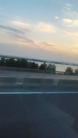 The video shows a view from a moving vehicle on a highway, likely during sunset. The road is visible in the foreground, with concrete barriers separating lanes. Beyond the barriers, a body of water is seen, with trees and distant hills on the horizon. The sky is a gradient of blue, pink, and orange, indicative of sunset. A truck and several cars are also visible on the highway. The motion blur suggests the video was filmed from a moving car.