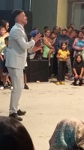 A man in a light gray suit, white shirt, and blue tie is standing in the center of an outdoor scene, holding blue juggling balls and appearing to perform. He is looking up and to his left, with a slight smile. A crowd of people, including men, women, and children, are gathered around him, watching the performance. Some of the children are in the front row, and others are further back. To the left of the performer, there is a large black speaker. To the right of the performer, a man wearing a dark blue t-shirt and light-colored shorts is holding a camera and filming the performance. The ground appears to be paved or made of concrete. The background is somewhat out of focus, but suggests buildings or structures. The overall atmosphere appears to be festive and engaging.