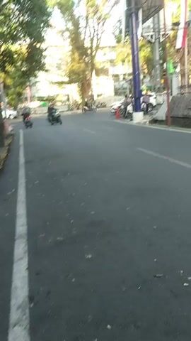 The video captures a street scene with a focus on a dark-colored motorcycle ridden by a person in black attire. The motorcycle is moving towards the right side of the frame. In the background, a white SUV is parked, and several other individuals are visible near a tall billboard structure. The environment appears to be urban, with trees and buildings lining the street. The lighting suggests it is daytime.