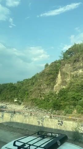 The video presents a daytime view of a hillside and a dry riverbed, seen from a vehicle moving across a bridge. The hillside is covered in vegetation, with a patch of bare earth visible. A small white building is situated near the riverbed. The riverbed itself is rocky and appears dry. The bridge has a concrete barrier and a metal railing. Above, the sky is blue with scattered white clouds.