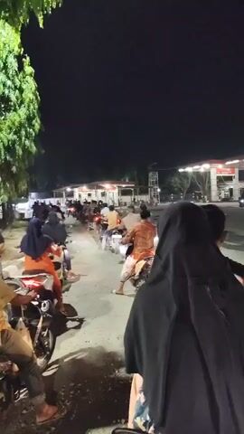 The video captures a nighttime scene at what appears to be a gas station. A long line of motorcycles is queuing up, with riders visible on many of them. In the foreground, a person wearing a black hijab is seated on a motorcycle, looking ahead. In the background, the gas station's illuminated canopy and pumps are visible. Several cars and other motorcycles are also present on the premises and on the road in front of the station. The overall lighting suggests it's dark, with artificial lights from the gas station and vehicles illuminating the area. The ground appears to be paved, with some wet patches or reflections visible, possibly from recent rain. The atmosphere seems to be one of waiting, as indicated by the queue.