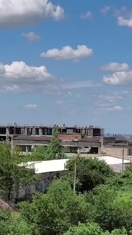 A long shot shows an unfinished concrete building under a bright blue sky with fluffy white clouds. The building appears to be multi-story and has exposed rebar on its upper level. Several figures, presumably construction workers, are visible on the top level of the building. Green trees and foliage surround the building, and some other smaller structures are visible in the background. The overall scene suggests an ongoing construction project in a semi-rural or industrial area.