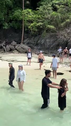 This media file captures a lively scene at Haad Hin Khom, Thailand. The image is a wide shot of a tropical beach with crystal-clear, shallow turquoise water in the foreground. A crowd of people, dressed in swimwear and casual beach attire, are visible on the sandy shore and wading in the water.

In the foreground, a man in a black t-shirt and dark shorts is interacting with a young woman in a black t-shirt and shorts. They appear to be looking at a smartphone. To their right, a woman in a white sundress and a hat is also looking at her phone, while a man in a light-colored t-shirt and blue shorts stands nearby. Further back, a mixed group of individuals, including men, women, and a child, are scattered across the beach. Some are walking, others are standing and observing, and a few are posing for photos. The beach is backed by a lush, green hillside covered in dense vegetation and rocky outcrops, suggesting a natural, unspoiled environment. A few scattered logs and driftwood are present on the sand.

The activity appears to be a casual beach outing or tour, possibly part of a vacation or excursion. The lighting suggests it's daytime, likely late morning or early afternoon, with bright sunlight casting shadows. The weather seems pleasant and warm, ideal for a beach visit. The presence of diverse groups of people and the relaxed atmosphere hint at a tourist destination. No visible text is present in the media file.
