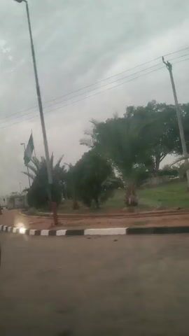 The video depicts an outdoor scene on a cloudy day. The camera is positioned within a moving vehicle, providing a view of the roadside. A Nigerian flag waves in the wind on the left side of the frame, planted in the center of a roundabout. In the background, the word "NIGERIA" is spelled out in large white letters, situated on a green grassy area. Two tall, dark signs flank the word. A black and white curb lines the edge of the roundabout. Trees and lamp posts are scattered around the perimeter. Power lines stretch across the overcast sky.