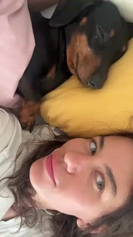 The video is a close-up selfie shot from a first-person perspective, angled downwards. A young woman with dark, curly hair and hazel eyes is in the foreground, looking up at the camera with a slight smile. Her lips are painted a soft pink. To her right, a black and tan Dachshund dog is sleeping soundly, its head resting on a yellow pillow. The dog's eyes are closed, and its paws are tucked beneath its chin. The background is blurred, but hints of a pink striped blanket and a light-colored garment, possibly a t-shirt, can be seen. The overall impression is one of intimacy and comfort.