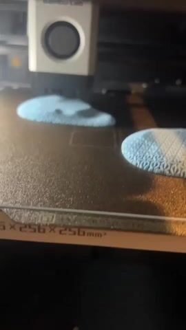 The video shows a close-up of a 3D printer bed with two blue objects being printed. The objects are round and appear to have intricate patterns on their surfaces, with one object showing a more defined geometric pattern than the other. A white 3D printer head is visible in the upper portion of the frame, moving over the objects. The printer bed is black and textured, with markings along the front edge indicating dimensions of 256x256x256 mm³. The lighting is dim, casting shadows on the printed objects.