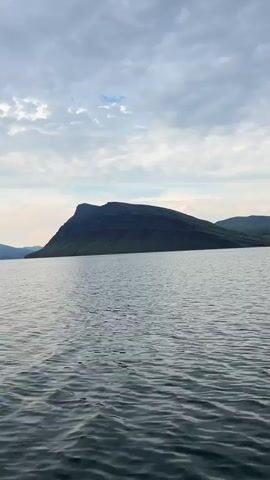 A boat is moving across a body of water towards a large, flat-topped mountain under a cloudy sky. The water is choppy with ripples. The mountain is dark green and appears to have layered rock formations. The sky is overcast with scattered clouds. The camera is mounted on the boat and is facing forward.
