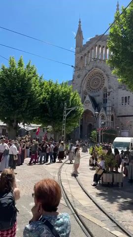 The video shows a crowd of people gathered in front of a large stone building with a tall tower. The building appears to be a church or cathedral. Many of the people are wearing historical costumes, and there are some modernly dressed people in the crowd as well. A street with cobblestone pavers and tram tracks runs through the scene. There are green trees lining the street, and the sky is bright blue and clear. A woman with short red hair is standing in the foreground, taking a photo of the scene. The loud sound of a horn can be heard in the background.