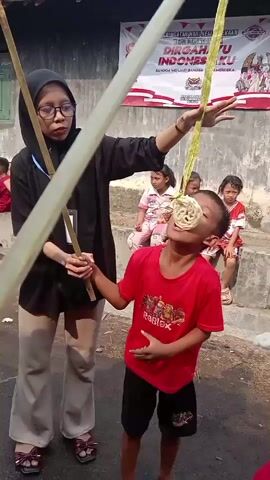 A group of children and at least two adults are gathered outdoors, likely for a celebration or event, possibly related to Indonesian Independence Day given the banner in the background. One adult, wearing glasses and a hijab, stands to the left holding a bamboo pole. A young boy in a red shirt is actively participating in a game, attempting to eat a cracker (kerupuk) that is suspended from a string attached to a bamboo pole. Another child, wearing an orange shirt, is also participating in the game, and another adult is visible on the far left, also holding a bamboo pole. Several other children are watching the game in the background. The environment appears to be somewhat rural, with buildings and a dirt or paved surface. The activity seems to be a fun, competitive game.