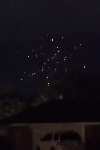 A night sky is visible above the roof of a house, with some trees in the foreground. Several bright lights are scattered across the sky, some appearing in clusters. One cluster of lights explodes outwards in a burst of light and color, resembling fireworks. Another bright light appears higher in the sky, possibly the moon. The scene is dark, suggesting it is nighttime.