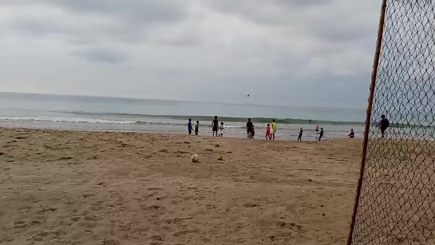 A group of people, some appearing to be children, are playing soccer on a sandy beach. The ocean with waves is visible in the background under a cloudy sky. A soccer net is positioned in the foreground, partially obscuring the view.