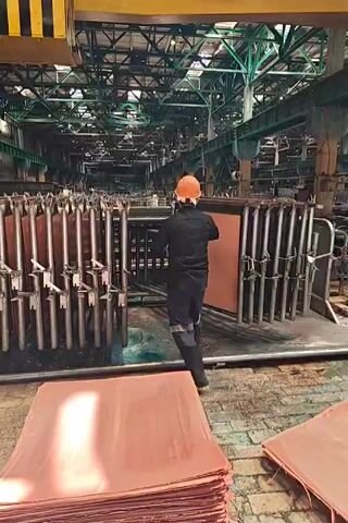 The video shows a worker in a factory setting, wearing an orange hard hat and dark work clothes. The worker is positioned between rows of what appear to be metal plates or sheets suspended in a liquid. The worker is seen manipulating one of the suspended plates, pulling it out of the liquid and then placing it back in. In the foreground, there are stacks of flat, copper-colored sheets. The environment appears to be a large industrial facility with high ceilings and exposed metal structures. The lighting is bright, suggesting artificial lighting within the factory. The actions suggest some sort of electroplating or refining process.