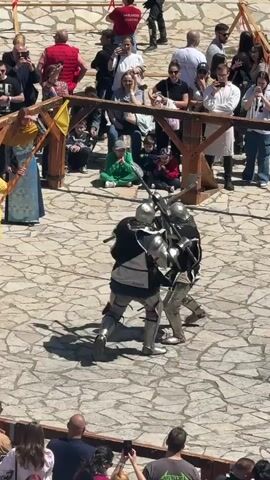 The video shows two knights engaged in a sword fight, surrounded by a crowd. The knights are wearing full suits of armor, and they are using their swords to strike at each other. The crowd is standing behind a wooden fence, watching the fight. The scene takes place on a stone-paved area. There are other people dressed in historical clothing. Some people in the crowd are taking photos with their phones. The lighting is bright and sunny.