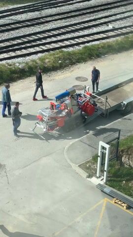 Four men stand around a small tracked vehicle loaded on a trailer. The vehicle is red, white, blue, and silver. One man is looking at it, two men are standing near the trailer, and one man is walking. Next to the trailer is a short fence, and in the background are railroad tracks.