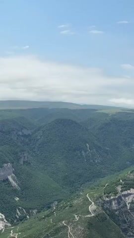 A scenic, wide shot of a lush green canyon under a blue sky with scattered clouds. The canyon walls are covered in dense vegetation, with patches of exposed rock and a winding road visible along one side. In the distance, rolling hills stretch towards the horizon. A bird of prey is seen soaring through the air in the upper right quadrant of the frame. The camera appears to be stationary or panning slowly.