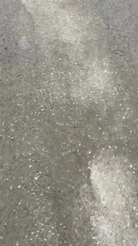 The video shows a ground level view of a paved or asphalt surface, then pans up and to the right to reveal a gray metal gate, a brown sliding door, and a hillside in the background. The ground is cracked and uneven in places. There is a dark-colored shoe visible at the bottom of the frame at the start of the video. The day appears overcast.