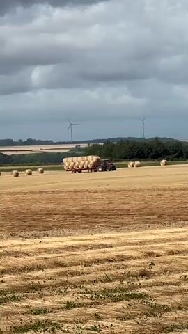 A red tractor pulls a trailer loaded with round bales of hay across a harvested field. The field is covered in straw with several round bales scattered throughout. In the background, a line of trees separates the field from a distant landscape with three wind turbines. The sky is overcast with dark, dramatic clouds.