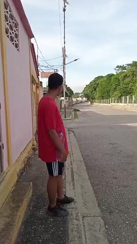 The video begins with a person standing on a sidewalk, facing away from the camera. The person is wearing a red t-shirt and black shorts with white stripes, and holding a clear plastic bottle. They are standing next to a light-colored building with a patterned wall. The sidewalk is next to a paved road, with a curb separating the two. In the background, there is a street with houses and trees. The camera then pans to the right, showing more of the street, a fence with pillars, and dense foliage. The video continues to pan, revealing a gated entrance with a covered structure and a small building to the right of the gate. The scene is outdoors, under a cloudy sky.