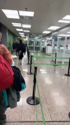 Here's a description of the video:

The video shows an indoor scene in what appears to be an airport or a similar transit facility.  Several individuals are lined up, navigating a queue guided by black posts connected with green straps. The people are walking toward a glass-enclosed area where personnel can be seen working behind desks. The person filming is also in the queue. The background shows a bright, sterile environment with fluorescent lighting fixtures in the ceiling.  There are signs with directions displayed above. The person recording appears to be carrying a red bag and a black backpack.