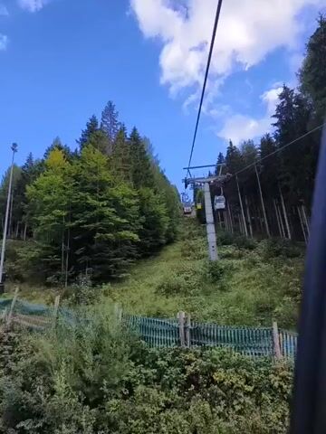 A ski lift ascends a grassy slope covered in trees. The sky is blue with scattered clouds. A few ski lift cabins are visible on the cable. The camera appears to be inside another ski lift cabin, looking upwards and forwards.