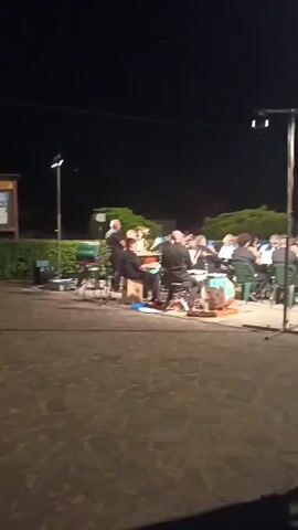 A group of musicians are performing outdoors at night. The scene is lit by artificial lights. In the foreground, there is a paved area. In the middle ground, a band is set up, with several individuals playing instruments such as drums and brass instruments. A conductor is visible at the front, directing the musicians. The musicians are seated on green chairs. Behind the band, there are trees and bushes. In the background, there is a building. The overall atmosphere suggests a live performance or concert.
