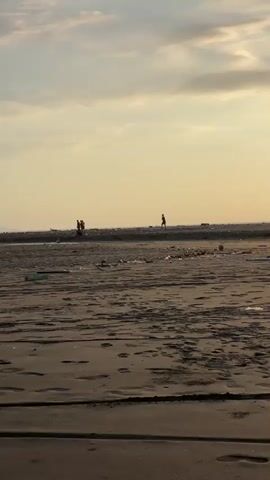 The video captures a wide shot of a beach scene at sunset. The sky is a blend of light yellow and pale blue with scattered clouds. In the midground, several people are visible on what appears to be a raised sandbar or an elevated part of the beach, some standing and others possibly sitting or stooping. One individual is walking on the right side of this elevated area, while a small group is gathered on the left. The foreground of the beach is sandy with noticeable ripples and footprints, suggesting it is either wet or recently exposed by the tide. Various small pieces of debris or trash are scattered across the sand in the midground, between the viewer and the people. The overall impression is a tranquil, somewhat distant view of beachgoers during the golden hour.
