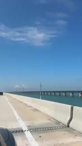 The video shows a car driving along a wide highway bridge. The sky is blue with scattered clouds. The bridge stretches far into the distance, with power poles spaced along the side. The water below is turquoise green. A car is visible ahead in the same lane. The concrete barrier lines the side of the road.