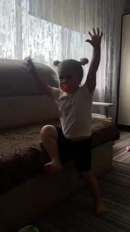 A young child with blonde hair and a pacifier is playing on a sofa. The child is wearing a white t-shirt and black shorts. The sofa is brown and has a patterned fabric. The scene appears to be indoors, with a window and curtains in the background. The light coming from the window suggests it is daytime. The child is actively moving, with one leg extended upwards and the other bent. The child's expression is not clearly visible. The overall mood seems playful and energetic. The location is identified as Peshnevo, Russia. No visible text is present in the media file.
