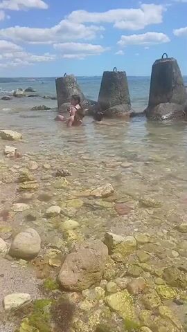 This media file captures a summer day at the beach in Osenovo, Bulgaria. The scene is set on a rocky shoreline with clear, shallow water. In the foreground, a young girl in a light-colored swimsuit stands on the sand, facing the water. Her posture suggests she is about to enter or is contemplating entering the sea.

Further into the water, near some large concrete breakwaters, two other individuals are visible. One person, in a dark swimsuit, appears to be reclining or sitting in the water. Another person is also in the water nearby, partially submerged. The breakwaters themselves are substantial, weathered concrete structures, indicating a man-made element within the natural beach environment, likely for coastal protection.

The water is transparent, revealing a bed of colorful rocks and pebbles beneath its surface. The beach is composed of sand and scattered stones of various sizes. In the background, a hazy coastline stretches across the horizon, suggesting a wide expanse of sea. The sky is bright blue, dotted with fluffy white clouds, indicative of fair weather and a sunny day. The overall atmosphere conveyed is one of leisure and enjoyment of a warm summer day by the sea. There is no visible text in the media file.
