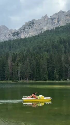A drone shot shows a bright yellow paddleboat with one person on a lake. The person is sitting and appears to be stretching or posing. The paddleboat is moving away from the drone. The lake is surrounded by a dense forest of pine trees. In the background, there are rugged, rocky mountains under a cloudy sky. The camera pans to the left, revealing a cluster of buildings on the shore of the lake, nestled among the trees, with more dramatic mountain peaks in the distance.