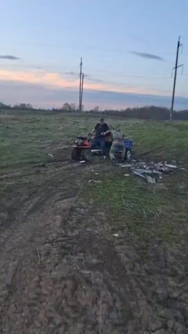 The video shows two men with a red ATV, one seated on it and the other pushing it from behind. The ATV has a blue trailer attached to it. The scene takes place in a muddy field with scattered debris. Power lines are visible in the background against a sky with a mix of blue and pink hues. The ground is uneven and appears recently traversed by the vehicle, leaving visible tracks in the mud. The man pushing the ATV is wearing camouflage clothing. The man on the ATV is wearing a dark shirt and appears to be steering. The environment suggests a rural or agricultural setting.