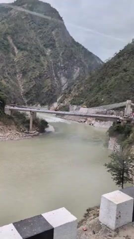 A wide, murky river flows through a valley surrounded by steep, green, and rocky mountains. A bridge spans the river in the middle distance, with cars visible on it. The mountains are dotted with small villages and terraced fields. The sky is overcast. The video appears to be filmed from a moving vehicle, possibly on a road running alongside the river.