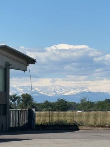 The video shows a view of snow-capped mountains from what appears to be an industrial or semi-rural area. The foreground features a low building with a corrugated metal roof and a metal fence, followed by a field with trees and greenery. Power lines stretch across the view, and a white car drives by on a road in the middle ground. A sign with the text "DIVIETO DI SCARICO" is visible near the road. The sky is blue with scattered white clouds, and the snow-covered mountains form a distant background.