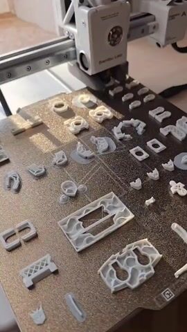 A 3D printer is in operation on a print bed. The print bed is covered with various small, white 3D printed objects. The 3D printer head is visible and appears to be actively printing or has just finished printing. The objects on the bed vary in shape and size, with some appearing to be intricate mechanical parts or decorative elements. The background is blurred but suggests an indoor environment.