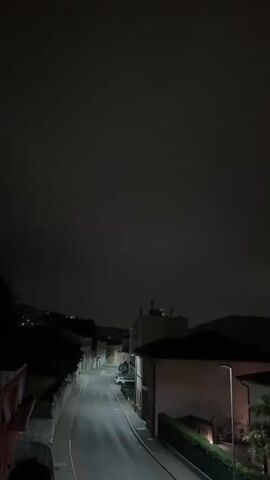A dimly lit, narrow, paved street winds uphill between buildings and trees at night. A single streetlamp illuminates the lower right portion of the street. The sky is dark and cloudy.