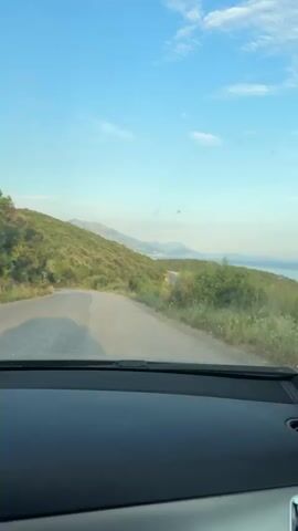 The video shows a view from inside a vehicle, looking out through the windshield and side window. The vehicle is driving on a paved road that curves to the left, uphill and to the left. The road is surrounded by lush green vegetation and trees. In the distance, a body of water, likely the sea, is visible. The sky is clear blue with a few scattered white clouds. The dashboard of the vehicle is visible in the foreground, and a side mirror reflects the passing scenery. The overall impression is that of a scenic drive through a coastal landscape on a clear day.