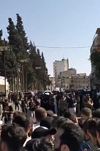 The video shows a large crowd of people, most of whom are facing the camera. In the background, there is a line of men in what appear to be black uniforms and vests. They are standing behind a barricade that runs along the street. Several vehicles are parked in the street behind the uniformed men. The crowd appears to be separated from the uniformed men by the barricade. Some of the people in the crowd are holding up a Syrian flag. The setting appears to be an urban area with buildings and trees. The weather appears to be sunny.