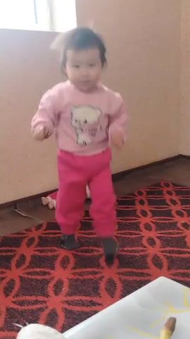 A toddler is the primary subject of this video. The child, dressed in a pink long-sleeved shirt and pink pants, is wearing dark socks. She is walking around the room, occasionally looking at the camera. Beside the child, on a red and black patterned rug, lies a doll dressed in similar pink clothing. The room features off-white walls, a window in the corner, and a table visible at the edge of the frame.